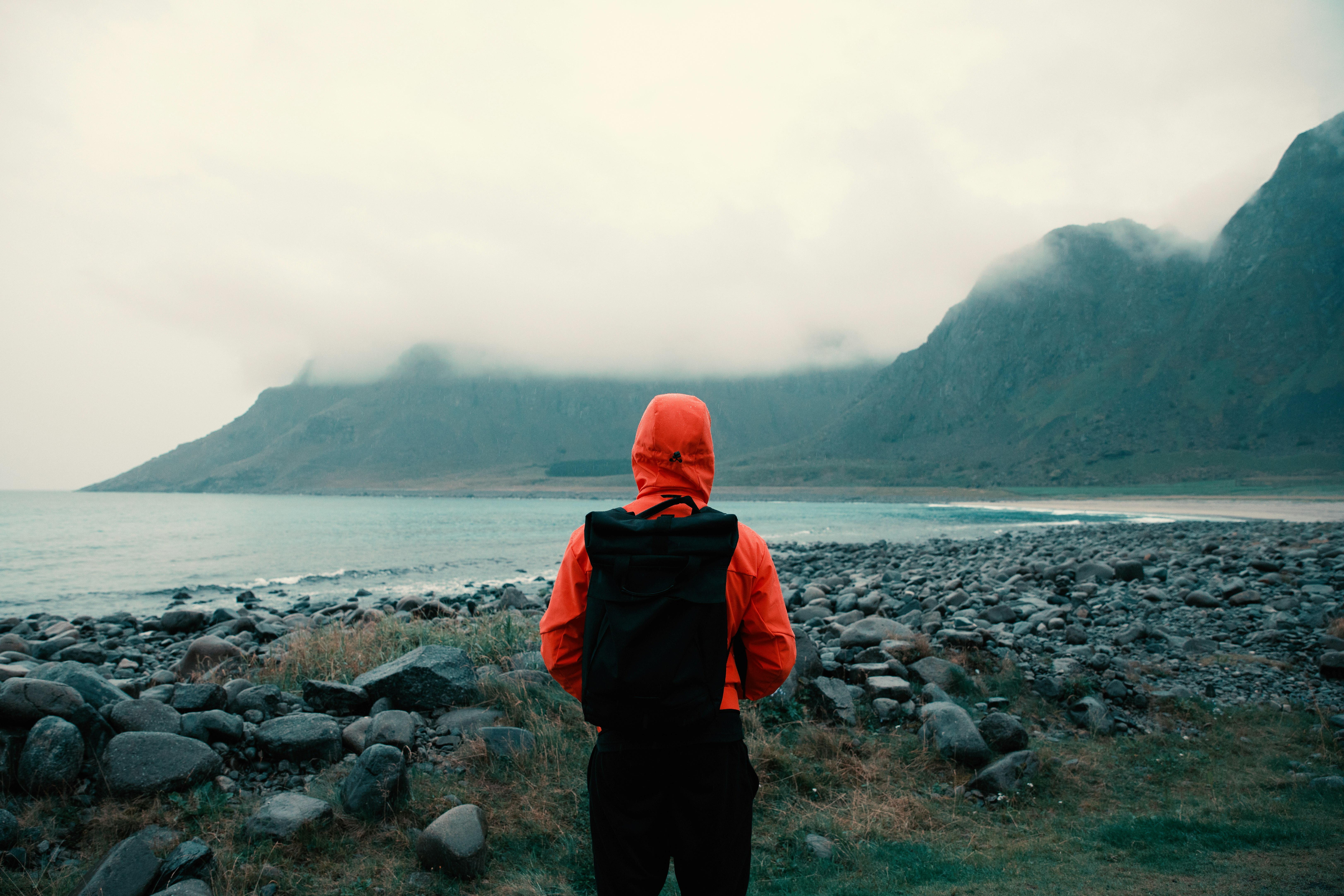 person in orange hooded jacket carrying bag looking at mountains coated with fogs